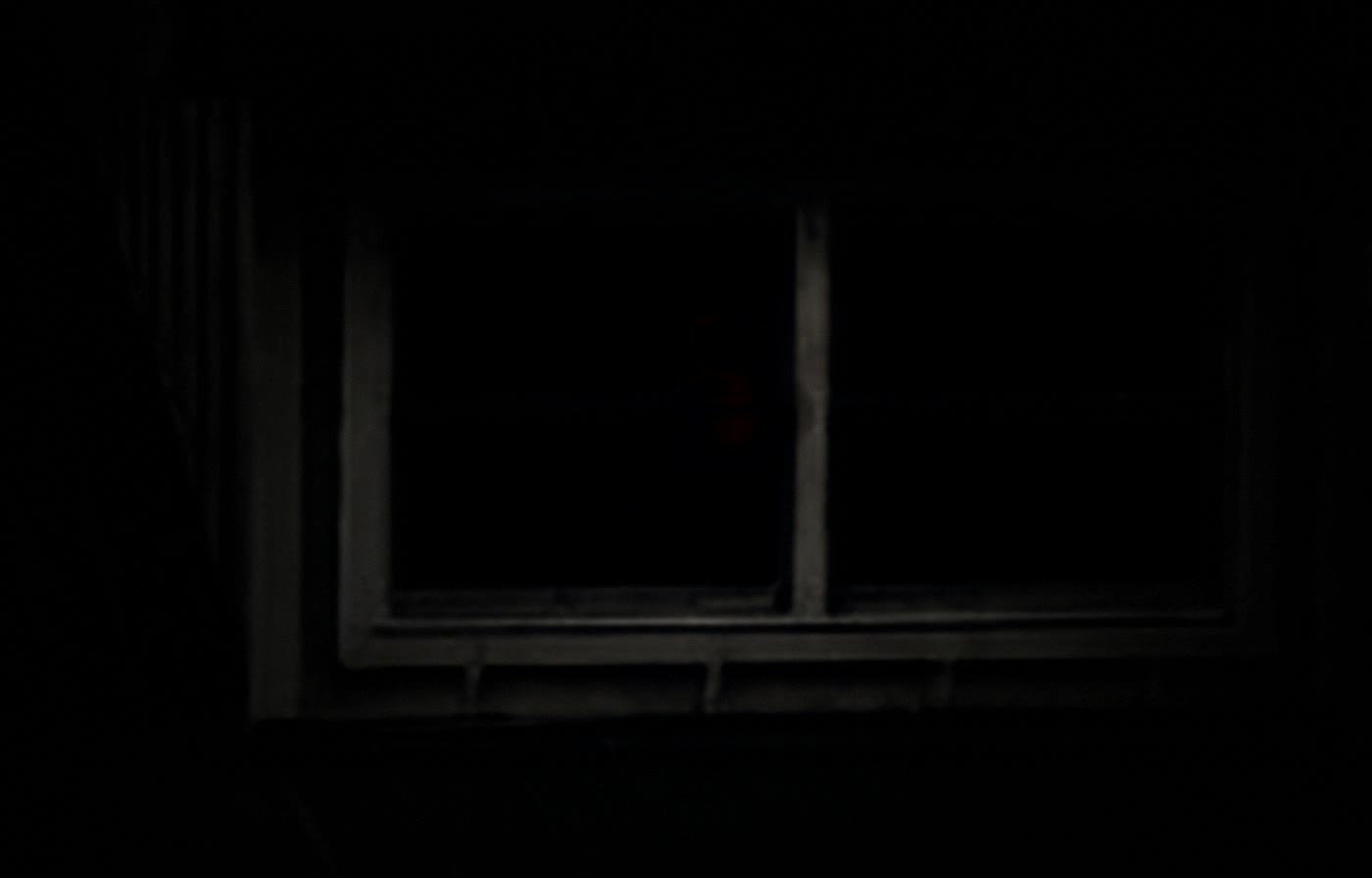 A dark upstairs window at night, dimly lit from somewhere inside the room.